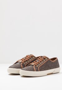 Braune Canvas-Sneaker mit strukturierter Oberfläche, beigefarbenen Akzenten, weißer Gummisohle, goldfarbenen Ösen und Schnürdesign mit dekorativem Markenlogo an der Seite.