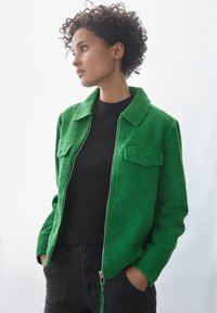 Grön texturerad jacka med dragkedja, två bröstfickor och kragdesign, kombinerad med en svart topp och mörka byxor.