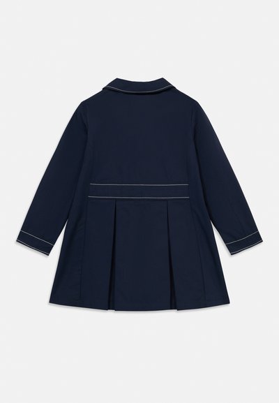 Μαντό σε ναυτικό μπλε με δομημένο σχεδιασμό, μακριά μανίκια και πλισέ πίσω μέρος. Διαθέτει αντίθετες λευκές ραφές κατά μήκος του γιακά και των μανσετών.