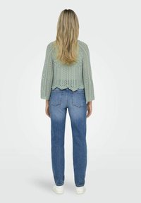 Pull vert clair en tricot avec un ourlet festonné, associé à un jean bleu taille haute et des baskets blanches. Cheveux longs et raides.