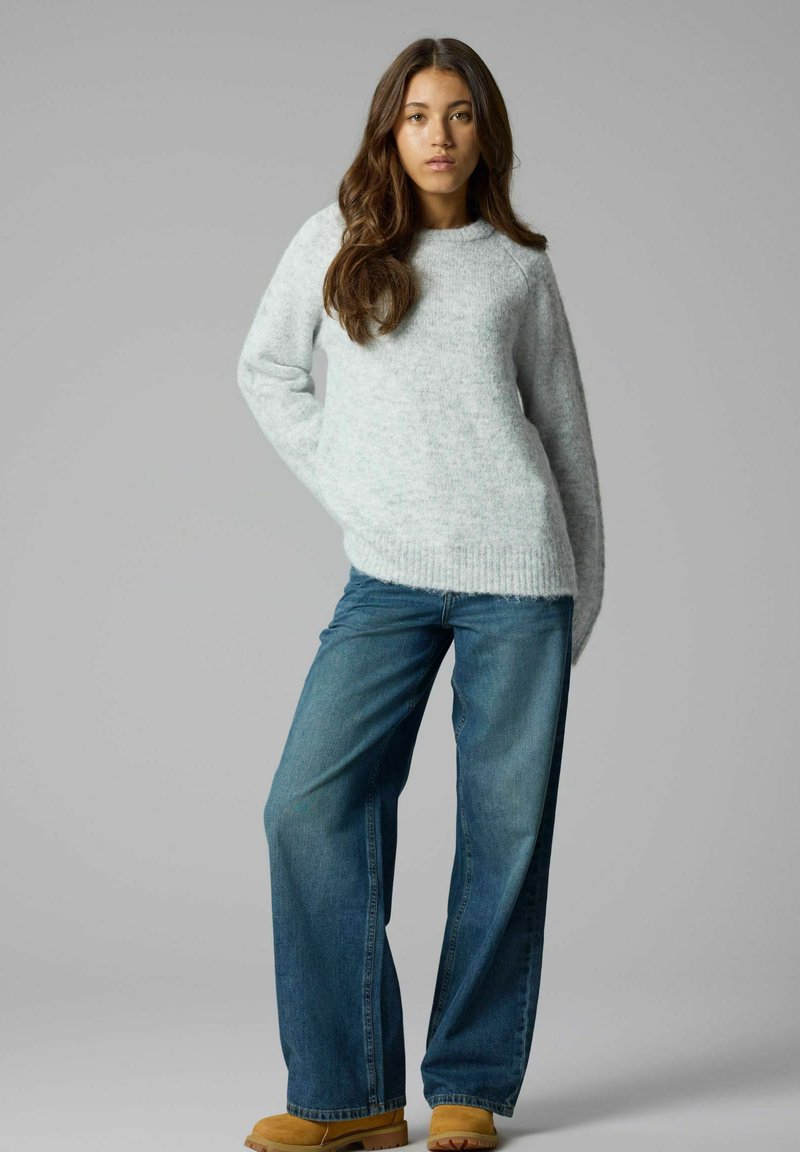 Jeune femme aux longs cheveux portant un pull gris clair, un jean bleu à jambes larges et des chaussures beige, debout devant un fond gris.