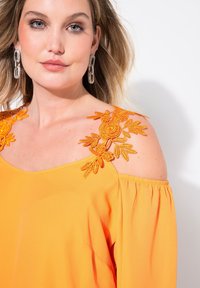 Orange Bluse mit schulterfreien Trägern und floralem Stickmuster in Gelb und Orange. Weicher Stoff mit lässiger Passform und elastischen Ärmeln.