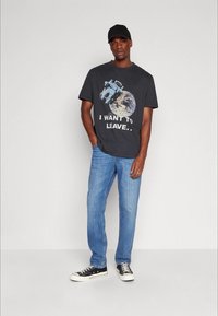 T-shirt gráfica cinza escuro com um design de astronauta e lua, combinado com jeans azul claro e ténis pretos. Um boné preto complementa o conjunto.