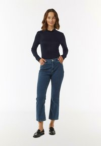 Navyblaues Langarmoberteil mit hohem Kragen, kombiniert mit hochgeschnittenen blauen Jeans mit Flared-Schnitt und schwarzen Loafers. Schlichtes, klassisches Design.