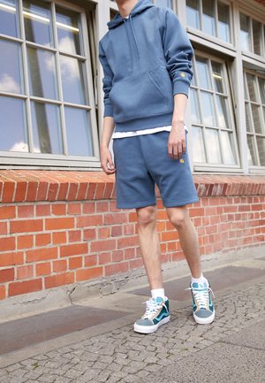 Blå hoodie og matchende shorts, lavet af bomuldsmateriale, loose fit, med lomme foran, gul logo detalje. Teal og hvide sneakers med teksturerede accenter.