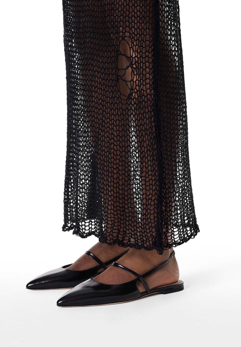 Ballerines noires à bout pointu en cuir verni, avec des fines lanières croisées. Le modèle porte un vêtement transparent en résille noire par-dessus.