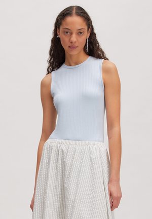 Femme aux longs cheveux bouclés portant un débardeur bleu clair côtelé et une jupe plissée blanche à fines rayures.