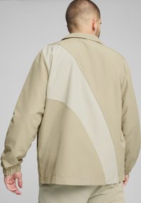 Beige jakke med en blød tekstur, der har et creamfarvet skråpanel på ryggen. Har elastiske manchetter og en afslappet pasform.