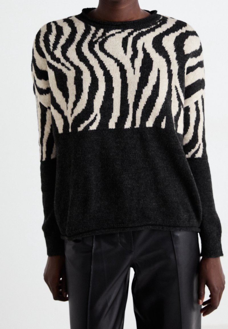 Personne portant un pull à motif zèbre noir et beige associé à un pantalon en cuir noir, debout devant un fond blanc uni.