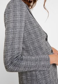 Grijze geruite blazer met een getailleerde pasvorm, voorzien van zwarte knopen, gestructureerde schouders en een soepele textuur.