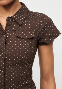 Καφέ κοντομάνικο πουκάμισο με μπεζ πουά, γιακά, κουμπιά με πατρόν και τσέπη με καπάκι στο στήθος, που φοριέται από ένα άτομο.