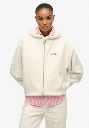 Femme portant un sweat à capuche crème zippé avec le texte "Superdry", superposé à une chemise rayée rose et blanche et un pantalon crème, debout les mains dans le dos.