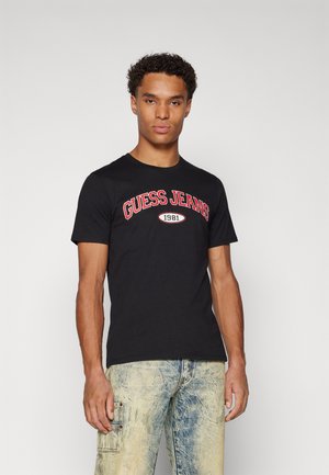 Maglietta nera in cotone con la scritta "GUESS JEANS" stampata in bold rosso su sfondo ovale bianco, abbinata a jeans chiari strappati.