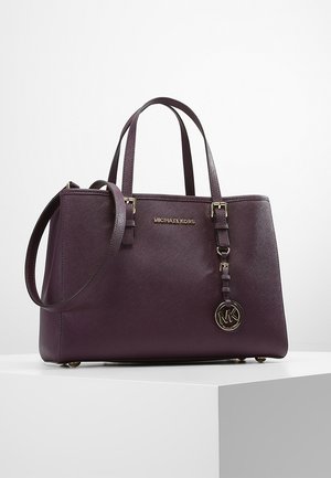 Donkerpaarse leren handtas van Michael Kors met dubbele handvatten, schouderriem en goudkleurige MK-logo hanger op een wit voetstuk.