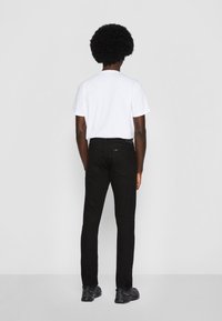 Persona con cabello rizado, vistiendo una camisa blanca de manga corta, jeans negros y zapatillas negras, de pie frente a un fondo liso de color gris claro.