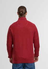 Mann mit braunen Haaren im Dutt, der einen roten Pullover und blaue Jeans trägt und mit dem Rücken zu einem schlichten grauen Hintergrund steht.
