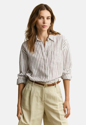 Femme aux cheveux bruns portant une chemise blanche à rayures verticales marron, un pantalon beige taille haute, et une ceinture marron, debout les mains détendues.