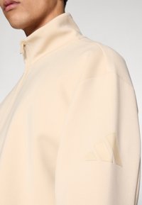 Ζακέτα με φερμουάρ σε ανοιχτό μπεζ ύφασμα, με ψηλό γιακά και υφασμάτινη υφή στο αριστερό μανίκι με σχεδιασμό λογοτύπου. Ομαλή υφή.