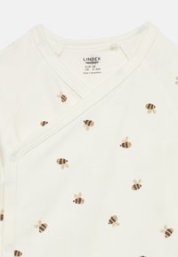 Άσπρο βαμβακερό μάρσιππος για μωρά με σχέδιο μέλισσας σε μπεζ και καφέ χρώματα. Κλείνει με κουμπάκια στο πλάι και έχει σχέδιο με V-λαιμόκοψη.