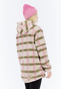 Persoon van achteren met een beige fleecejack met een groen en roze ruitpatroon en een roze-wit gestreepte gebreide muts.