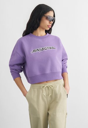 Žena v fialovej crop mikine s grafickým textom, béžových nohaviciach so šnúrkou a tmavých slnečných okuliaroch na zaviazanie, pózujúca s rukami v vreckách.