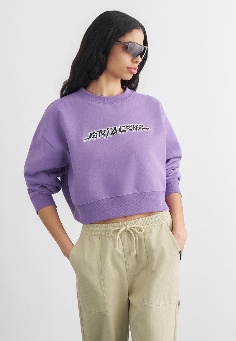 Naine, kes kannab lilla lühendatud dressipluusi graafilise tekstiga, beeže paelaga pükse ja tumedaid ümber pea mähitud päikeseprille, poseerides käed taskutes.