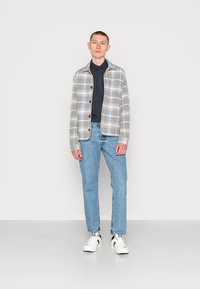 Camisa de padrão xadrez em cinza claro e bege, combinada com uma camisa azul escura, jeans azuis e ténis brancos com detalhes pretos.