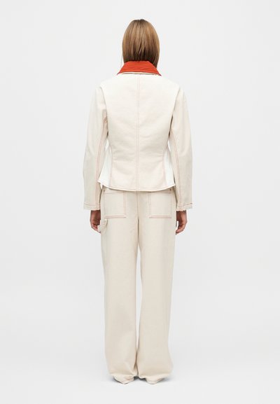Κρεμ σακάκι με πορτοκαλί γιακά, εφαρμοστή γραμμή και εμφανείς ραφές, συνδυασμένο με ευρύχωρο κρεμ παντελόνι με τσέπες.