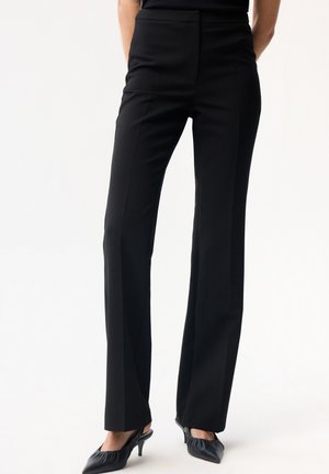 Person trägt hoch taillierte, gerade geschnittene schwarze Hose mit einer Bügelfalte vorne, kombiniert mit schwarzen spitzen Absätzen mit gerüschtem Detail.