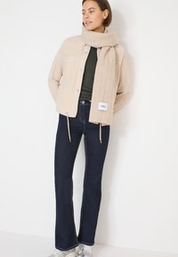 Beige kordfløjlsjakke med lommer, grøn top, mørke vidde jeans og et lysebrunt mønstret tørklæde med en label, der siger "CECI."