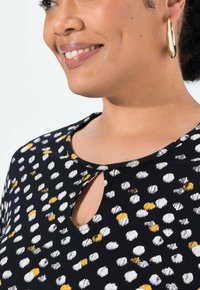 Top nero con scollo a chiave, caratterizzato da un motivo di punti bianchi e gialli. Realizzato in tessuto morbido, ha un colletto rotondo e maniche corte.