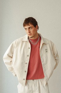 Veste boutonnée beige clair sur une chemise rose, colliers en chaîne argentés, manches courtes et texte brodé sur la veste. Design minimaliste.