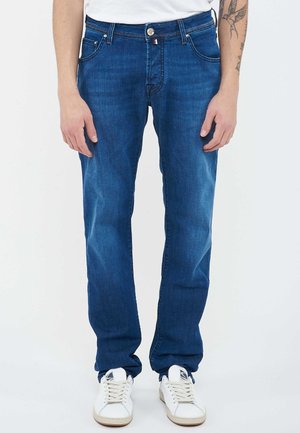 Man wearing blue jeans and white sneakers, standing with arms relaxed at sides, showing a tattoo on left forearm.