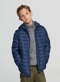 Giubbotto imbottito blu navy con cappuccio, caratterizzato da una texture trapuntata e chiusura frontale con zip. Indossato sopra a un maglione grigio a trecce e pantaloni verde oliva.
