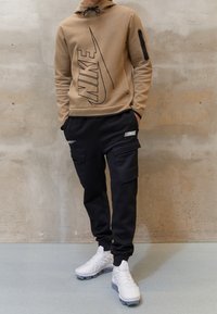 Nike beige Kapuzenpullover mit großem schwarzem Logo, lässige schwarze Cargo-Hosen mit doppelten Seitentaschen und weißen Sportschuhen mit strukturierten Details.