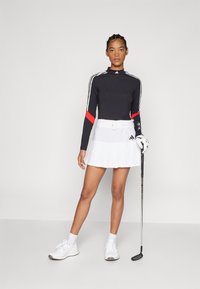 Μαύρη μπλούζα με μακριά μανίκια, κόκκινες λεπτομέρειες και λευκό σχέδιο με τρείς ρίγες, συνδυασμένη με λευκή πλισέ φούστα γκολφ και λευκά αθλητικά παπούτσια. Κρατάει ένα μπαστούνι γκολφ.