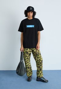 Camiseta negra con el logo azul de "Carhartt", pantalones verdes con patrón de camuflaje, bolso de hombro negro texturizado y zapatillas negras con detalles verdes.