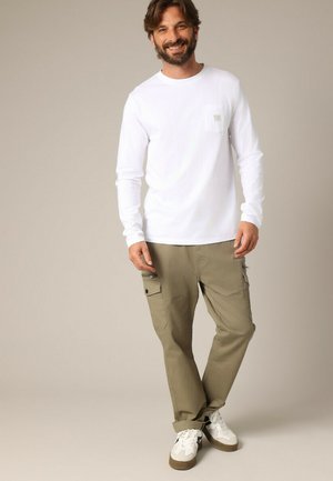 Witte longsleeve shirt met een klein borstzakje, gecombineerd met olijfgroene cargobroeken met meerdere zakken en strakke witte sneakers.