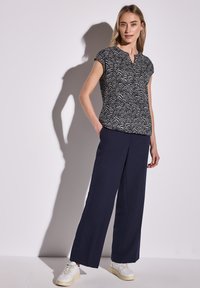 Blusa blu navy a fantasia con maniche corte e scollatura a chiave, abbinata a pantaloni larghi blu navy e sneaker bianche. Tessuto con una texture liscia.