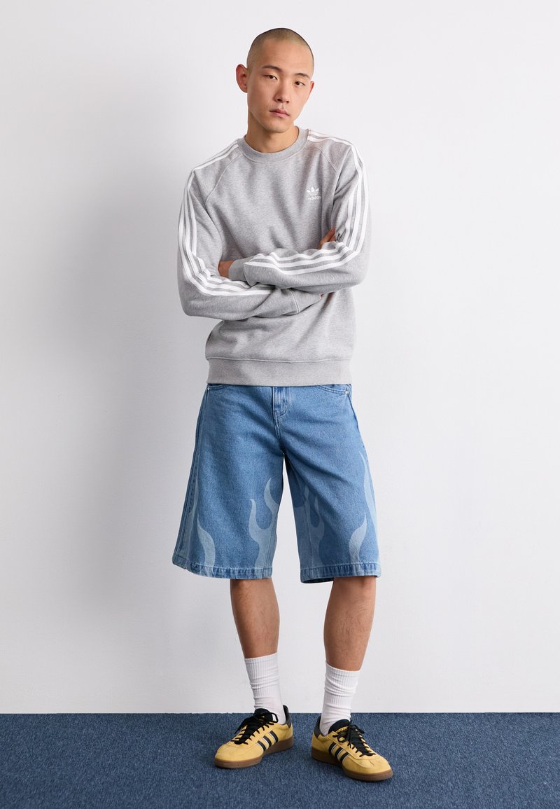 Sudadera gris con rayas blancas, pantalones cortos de mezclilla azul con diseño de llamas y zapatillas amarillas con detalles negros, contra un fondo liso.