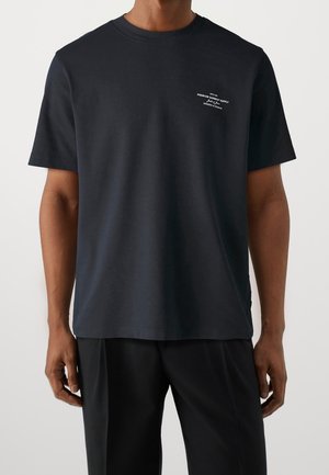 Mężczyzna ubrany w czarną koszulkę z krótkim rękawem z małym białym napisem na lewym górnym ramieniu i czarne spodnie, stojący na jasnym tle.