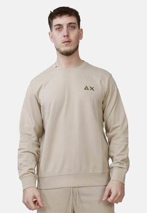 Ung mand iført beige sweatshirt med lille grøn trekant og X-logo, stående foran ensfarvet hvid baggrund.