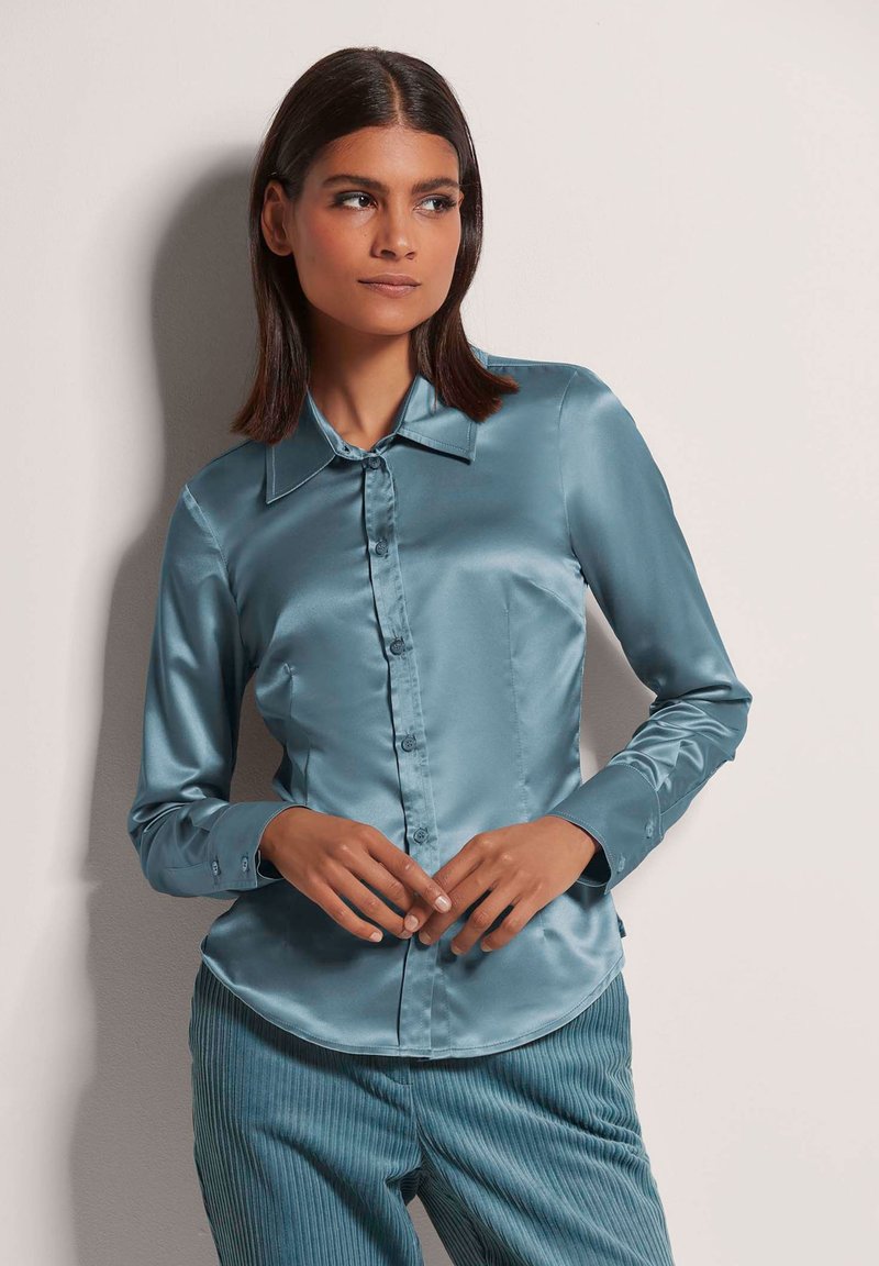 Langarmhemd mit Kragen aus teal Satin, mit knöpfbarem Frontverschluss und glatter Textur. Das Model posiert mit Händen vor einem hellen Hintergrund.