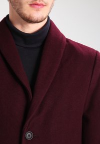 Bordeauxroter Wollmischmantel mit einem Reverskragen, ausgestattet mit einem Einzelknopfverschluss und strukturierter Stoffoberfläche, kombiniert mit einem schwarzen Rollkragenpullover.