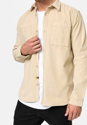 Man draagt een beige corduroy overhemd over een wit T-shirt en zwarte broek, hij stelt de kraag van het overhemd bij met zijn linkerhand.