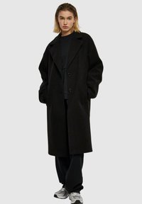 Sort, lang modell frakk med notched krage, dobbelspent knapp, og to frontlommer. Båret over en svart topp og avslappede bukser.
