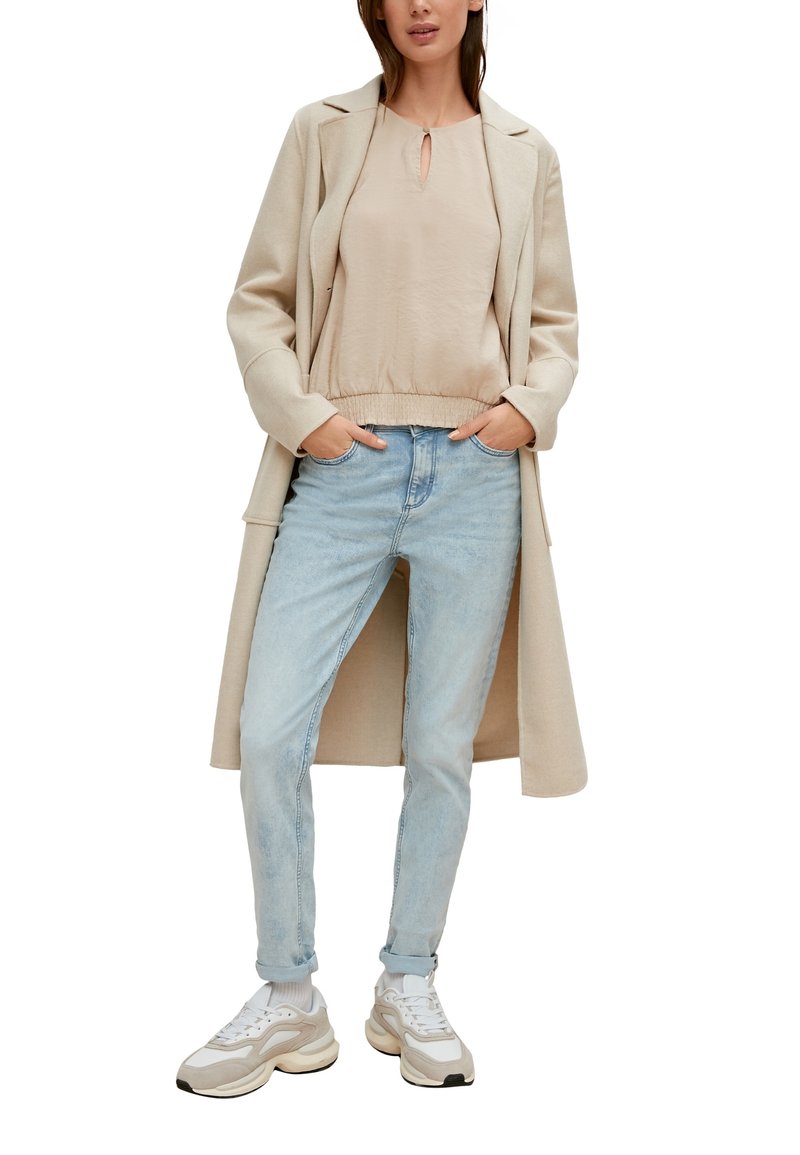 Femme portant un long manteau beige, un blouse beige, un jean bleu clair et des baskets blanches, se tenant les mains dans les poches sur un fond blanc.
