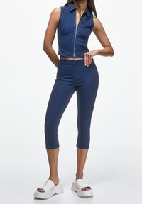 Kvinna klädd i ärmlös blå zip-up topp och matchande cropped byxor, stående med ena handen på höften och vita plattformssandaler.