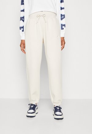Cream-colored joggers with an elastic waistband and drawstring. Ribbed cuffs at the ankles, paired with navy and white sneakers.