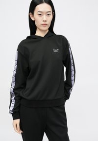 Čierna mikina s kapucňou s logom na prednej strane a purpurovými znakmi pozdĺž rukávov. Má voľný strih a mäkkú textúru, doplnená o nohavice v rovnakej farbe.